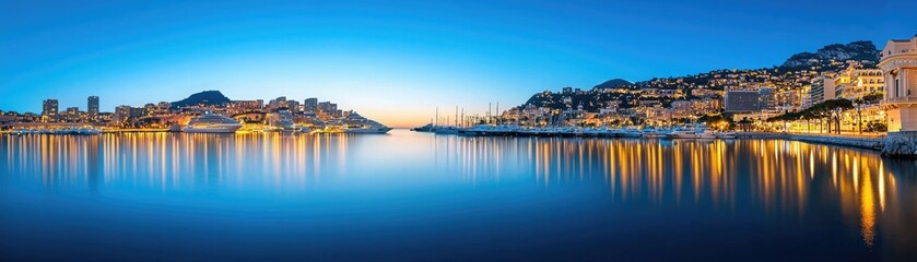 Fototapeta premium A serene panoramic view of a waterfront city at twilight, reflecting lights on calm waters, with buildings lining the shore.