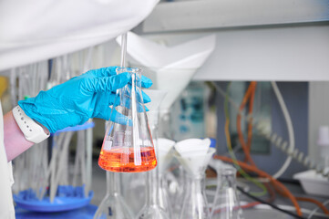 A scientist in a lab wearing gloves holds a flask with orange liquid during an experiment in a...