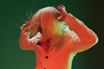 A person with long, light colored hair, dressed in a stylish blazer, poses dramatically against a...