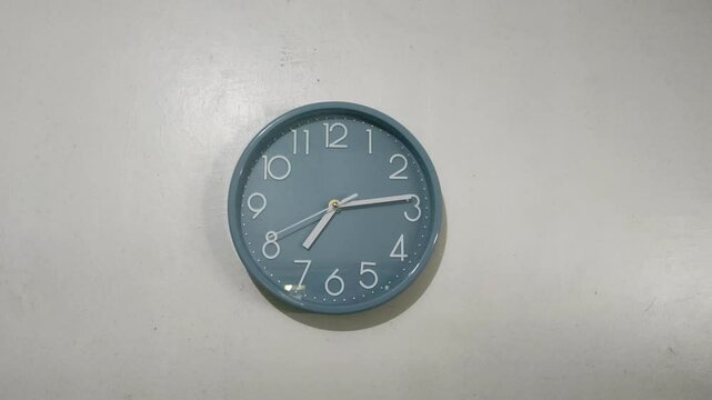 A blue wall clock shows the time as a little past 7. The simple design features white numbers and hands against a plain background, creating a minimalist appearance.