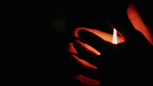 Woman hands touching the fire of a candle on dark black background, blur lights behind. Female holding candle with fire flame with empty space. Magic ritual with one candle wax fire. Close-up emotion 