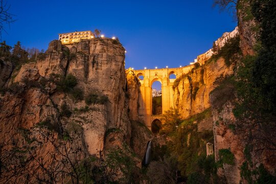 Scenic view of Puente Nuevo bridge in Ronda, Spain, illuminated at dusk with a vibrant blue sky.