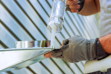 Construction worker uses caulking gun to apply sealant to ventilation metal flashing surface, ensuring it is weatherproofed.