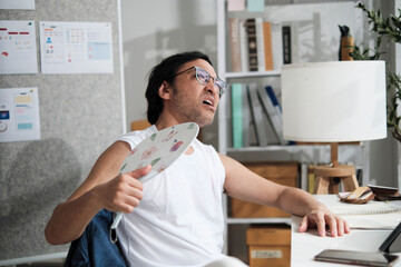 A male Asian worker is unhappy working with hot temperatures, taking off with vest at casual office...