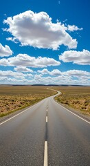 Naklejka premium A winding asphalt road stretches across a vast open countryside landscape with a bright blue sky and fluffy white clouds above The wide angle perspective creates a scene