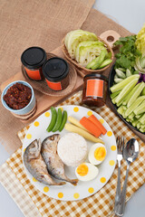 A meal setup featuring a cup of thai chili paste, a plate with two pieces of cooked fish, white rice, boiled egg and vegetables on  wooden tray