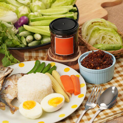 A meal setup featuring a cup of thai chili paste, a plate with two pieces of cooked fish, white rice, boiled egg and vegetables on  wooden tray