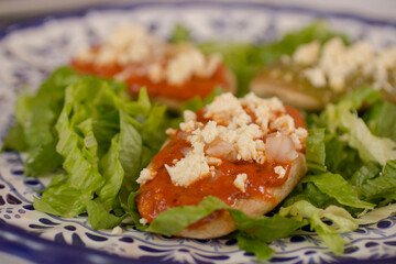 Close-up of a traditional Mexican tlacoyo with chipotle sauce, fresh cheese, diced onions, and shredded lettuce on a colorful Talavera plate.