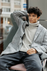 Fashionable young man sitting in front of a window, wearing a grey sweater, black turtleneck, and jeans