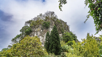 Obraz premium A picturesque mountain against the blue sky and clouds. There is green vegetation on the steep slopes and the top. Lush thickets of trees in the foreground. Fubo Hill. Guilin. China.
