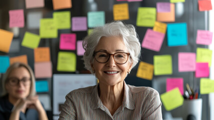 Smiling elderly woman with glasses in creative office environment, surrounded by colorful sticky notes, exuding sense of positivity and collaboration