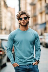 A stylish young man stands confidently on a city street, wearing a light blue color long sleeve T-shirt mockup and sunglasses. He has wavy hair and a casual yet fashionable look.