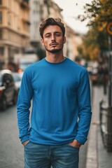 A young man stands confidently on a city street, wearing a blue color long sleeve T-shirt mockup and jeans. He has a stylish haircut and a serious expression, with cars and trees in the background.