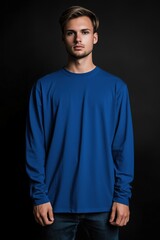 Obraz premium A young man stands confidently in a blue color long sleeve T-shirt mockup against a dark background. He has a neutral expression and is looking directly at the camera.