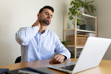 Man suffering from neck pain after working with laptop. Male self massaging neck and shoulders at...