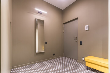 Spacious entrance hall, decorated in beige. Floor tiles with a pattern. A yellow banquette .and a mirror on the wall.