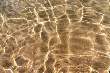 Through the transparent water of the ocean or sea you can see the sandy bottom with ridges of sand. Sun glare and ripples. View from above