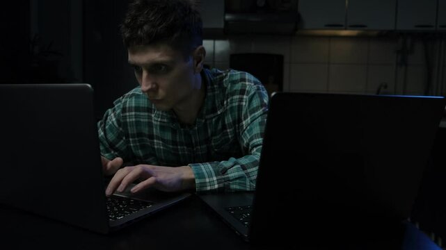 Focused young man working late at night on two laptops in a dimly lit room, intensely concentrating on his tasks. Dual screens emphasize productivity, technology, and multitasking.