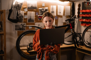 Cute blonde woman using a laptop computer in bicycle repair shop. Running a business and start-up