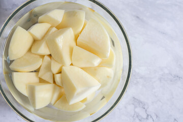 sliced potato soaking in a glass bowl of water. On a white marble background