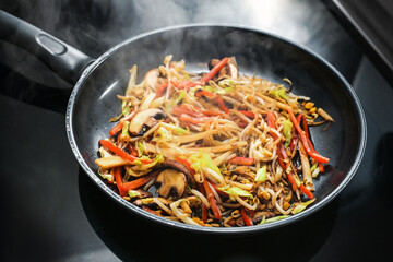 Steaming vegetables stir fried in a black pan, such as mushrooms, red bell peppers, soybean sprouts and cabbage for cooking an Asian meal