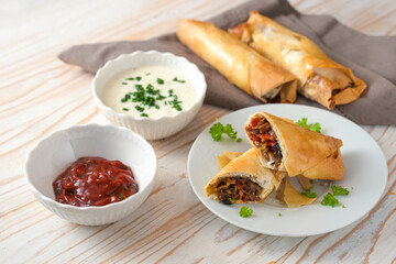 Crispy spring rolls from filo pastry stuffed with spicy vegetables and two dips on a light wooden table, homemade finger food in Asian style, selected focus