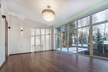 An empty room in a country house. Parquet, luxurious chandelier, unfurnished. Panoramic window with a view of the winter landscape.