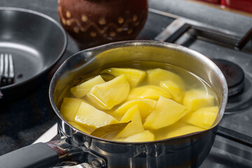 Boiled potatoes in saucepan in water with bay leaf. View on a stainless steel gas cooktop on a black table with red ceramic tiles on the wall close-up.