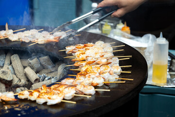Grilling skewers of shrimp on a large circular grill at a vibrant street food market during a sunny day.