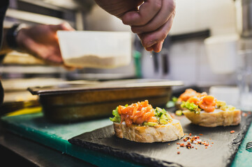 A close-up of a chef preparing bruschetta, topping toasted bread with fresh ingredients. Hands sprinkle seasoning over the dish, showcasing the art of culinary presentation in a kitchen setting.