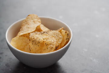 White bowl with organic potato chips sprinkled with fresh ground black pepper on concrete background with copy space