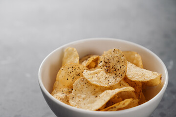 White bowl with organic potato chips sprinkled with fresh ground black pepper on concrete background with copy space