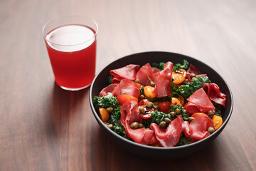 Salad with kale, bresaola and cherry tomatoes decorated with capers in black bowl and glass of red drink on walnut wood table