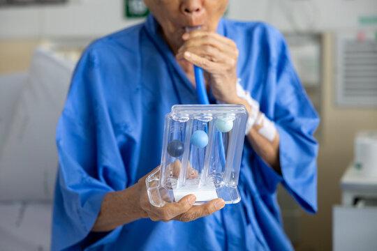 Senior asian man using incentive spirometer device for breathing exercise rehabilitation to recover from lung cancer after surgery due to heavy smoking in hospital bed