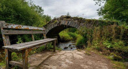 Fototapeta premium Old wooden bench by stone bridge over small stream in lush, green forest setting