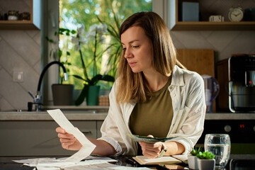 Stressed woman reviewing bills at home. Female organizing receipts, sitting at kitchen table with...