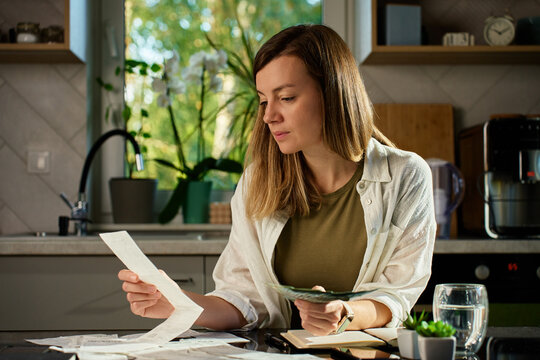 Stressed woman reviewing bills at home. Female organizing receipts, sitting at kitchen table with laptop and paper documents. Concept of budgeting and managing personal finances