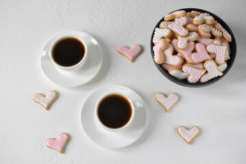 Top view of two espresso cups and heart-shaped cookies with pink icing in a black deep bowl. White...