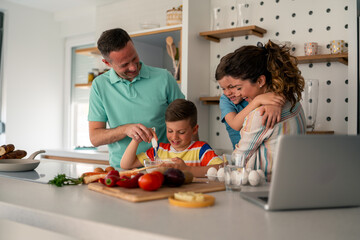 Caucasian family in casual attire teaches cooking skills to children while following digital recipe in modern domestic kitchen with wooden accents and organized storage.