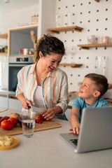 Caucasian mother and young son share cooking lesson in modern kitchen, following digital recipe while chopping ingredients on wooden cutting board near laptop.