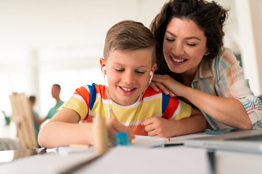 Caucasian mother in casual attire assists elementary-aged son with homework assignment using pencil and notebook at bright home study space.
