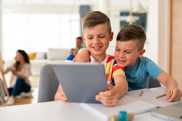 Two Caucasian brothers in casual attire explore educational content on a digital tablet while sitting at dining room table in home setting.