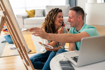 Obraz premium Mid adult Caucasian couple in casual attire creates artwork using paint supplies, sharing genuine laughter during their creative activity in comfortable living room setting.