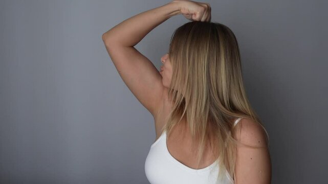 Young smiling cheerful strong fit blonde woman raising arm and showing bicep kissing her muscles on a dark grey background. Feminism, girl power, equal women's rights, independence, sports concept