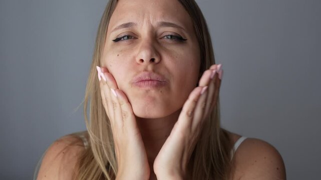 Young blonde woman massaging her face quickly slapping hard herself on the cheeks to cheer up, to improve blood circulation, relax the muscles, tighten skin on a dark grey background