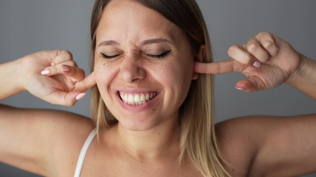 A young smiling caucasian blonde woman covers her ears with her fingers putting ear plugs squinting eyes. The girl doesn't want to hear the noise