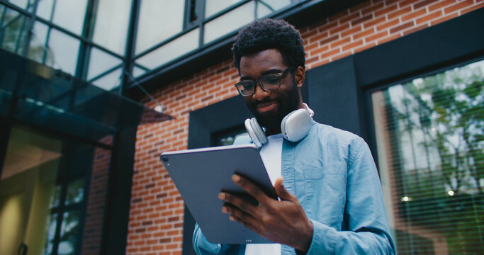 Camera moving closer to African American man standing outside under modern building. Using large tablet device. Typing on screen with one hand. Wearing headphones on his neck. Smiling.