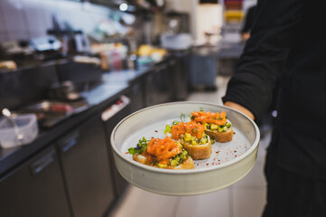 A chef holds a plate of vibrant appetizers in a bustling kitchen. The dish features small toasts topped with fresh ingredients, showcasing culinary creativity and attention to detail.