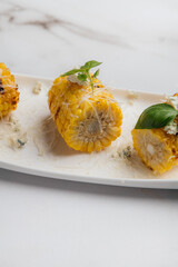 Three pieces of grilled corn on the cob are served on a white rectangular plate, topped with blue cheese, basil leaves, and grated cheese. Restaurant or cafe menu, cookbook recipe