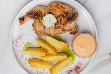 Top view of a delicious grilled chicken served with potato wedges, a creamy sauce, and herbs on a white plate, creating an enticing culinary scene. Restaurant or cafe menu, cookbook recipe